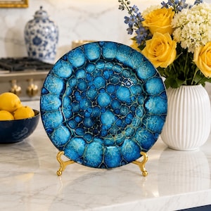May include: A round decorative plate featuring a vibrant blue and teal cell-like pattern with a glossy finish. The plate is displayed upright on a small gold-toned metal stand, showcasing a complex, organic design of interconnected circular shapes.