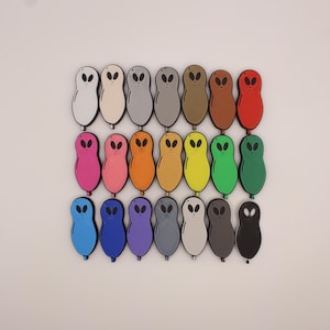 Puede incluir: Una colección de veinte imanes con forma de alienígena en varios colores, incluyendo blanco, rosa, naranja, amarillo, verde, azul, morado, gris, marrón, rojo y negro. Cada imán tiene un diseño minimalista con dos ojos ovalados.