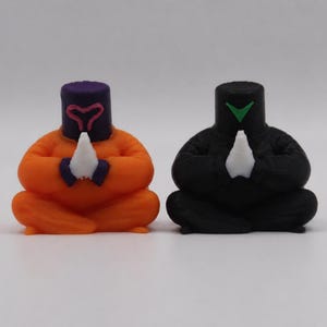 May include: Two small, stylized figurines in a seated pose. One is orange with a purple head and a pink heart-shaped design. The other is black with a green triangle on its head. Both have white hands clasped in prayer.