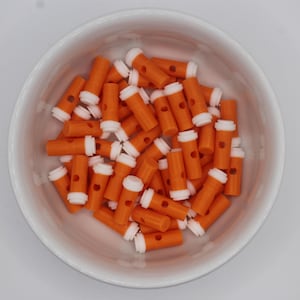 May include: A white bowl filled with numerous orange and white lancets. The lancets are cylindrical with a white cap and a small hole. The bowl is on a white surface.