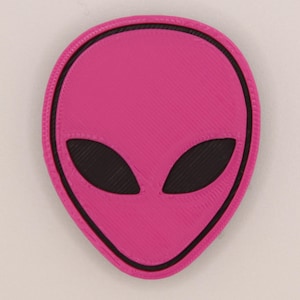 May include: A pink alien head patch with a black outline and black eyes. The patch has a textured surface.