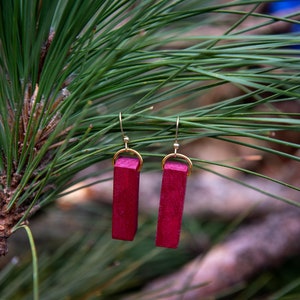 May include: A pair of earrings with a burgundy wood rectangular pendant and gold-toned hooks. The earrings are hanging from a pine tree branch.