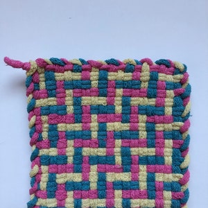 May include: A woven pot holder with a pink, yellow, and blue checkered pattern. The pot holder has a braided edge.