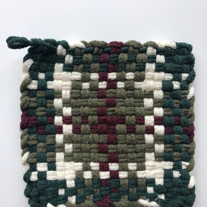Puede incluir: Un posavasos tejido con un patrón de cuadros verdes, blancos y burdeos. El posavasos tiene un lazo para colgar.