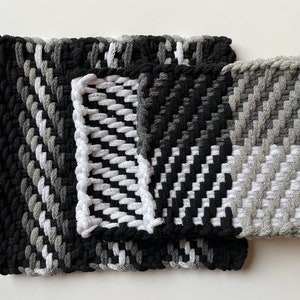 May include: A set of three woven pot holders in black, white and grey. The pot holders are made of a thick, textured fabric and have a simple, geometric design.