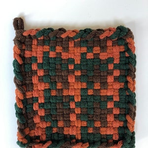 May include: A woven pot holder with a brown, green and orange chequered pattern. The pot holder has a fringe border.