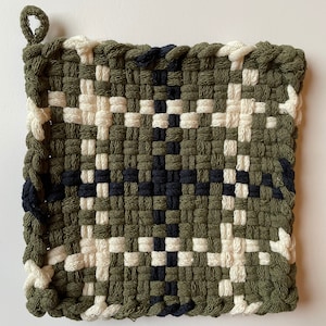 May include: A woven pot holder with a green, white and black chequered pattern. The pot holder has a loop for hanging.