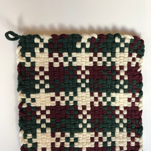 May include: A square woven pot holder with a loop handle. The pot holder is made with a green, burgundy, and cream coloured yarn.