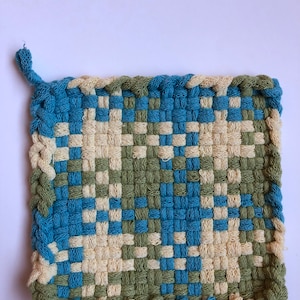 May include: A woven pot holder with a blue, green, and cream checkered pattern. The pot holder is made of cotton and has a fringed edge.