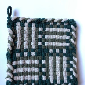 Puede incluir: Salvamanteles cuadrado tejido en tonos verdes y beige. El salvamanteles presenta un diseño tejido con un borde trenzado y un lazo para colgar. La paleta de colores incluye verde oscuro, verde oliva y beige.