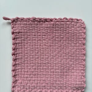 May include: A square, woven, pink potholder with a loop for hanging. The potholder is made of a tightly woven material, with a textured surface and a decorative border. The color is a soft, muted pink.