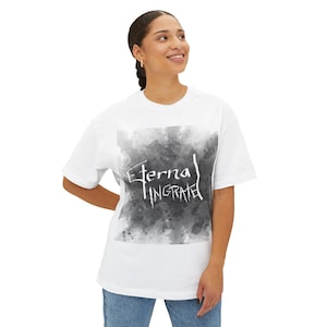 May include: White t-shirt featuring the text "Eternal Ingrate" in a stylized font. The text is set against a grayscale watercolor-style background. The shirt is a casual, oversized fit.