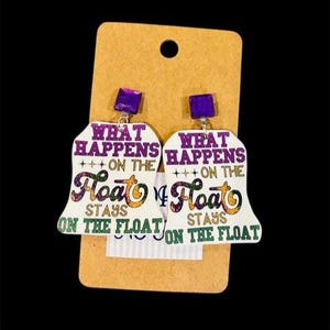 May include: A pair of white earrings with a bell shape, featuring the text "WHAT HAPPENS ON THE FLOAT STAYS ON THE FLOAT" in purple, orange, and green. The earrings have purple square studs and are displayed on a tan card.