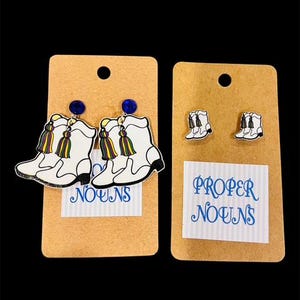 May include: Two pairs of earrings on tan cards. One pair features white cowboy boot designs with colorful tassels and blue gem studs. The other pair has smaller white cowboy boot designs. The text "Proper Nouns" is on one card.