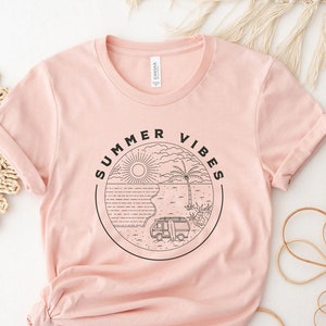 Puede incluir: Una camiseta rosa claro con las mangas enrolladas. La camiseta presenta un gráfico circular con las palabras "SUMMER VIBES" alrededor de una escena de playa con una furgoneta, una tabla de surf, una palmera y el sol. La camiseta es de material suave.
