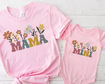 Könnte beinhalten: Rosa T-Shirt mit dem Wort "MAMA" in bunten Buchstaben mit Blumen und Blättern. Ein passender rosa Baby-Strampler mit dem Wort "MINI" in bunten Buchstaben mit Blumen und Blättern.