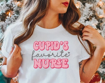 以下が含まれることがあります： ピンクとグレーの文字で「CUPID'S favorite NURSE」と書かれた白いTシャツです。