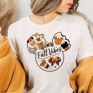May include: A cream-coloured t-shirt with a Mickey Mouse head design, incorporating autumn-themed graphics. The design includes pumpkins, cupcakes, and the text "Fall Vibes". The person is wearing a mustard-coloured sweater.