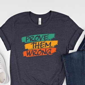 May include: A dark blue t-shirt with the words "PROVE THEM WRONG" in colorful block letters. The letters are outlined in black and have a paint-splattered effect. The shirt is laid flat on a white surface with a pair of white sneakers and blue jeans.