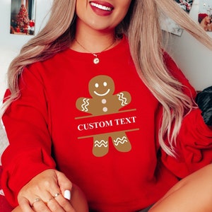 May include: A red sweatshirt with a gingerbread man graphic and the text "CUSTOM TEXT" printed on the front. The gingerbread man is brown with white icing and a smiling face.