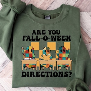 May include: Green sweatshirt with the text "ARE YOU FALL-O-WEEN DIRECTIONS?". The design features three cartoon ghosts at desks with books and an apple. The ghosts are made of colorful patchwork.