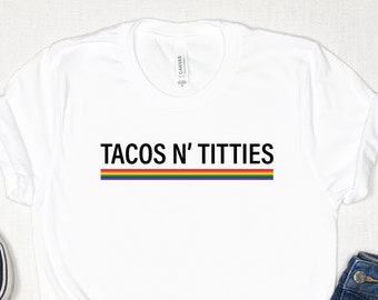 Könnte beinhalten: Weißes T-Shirt mit dem Text "TACOS N' TITTIES" in schwarzer Schrift mit einem Regenbogenstreifen darunter. Das Shirt liegt flach auf einer weißen Oberfläche mit einem Paar schwarzer Turnschuhe und einer blauen Jeans.