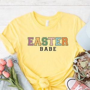 Może przedstawiać: Żółta koszulka z napisem "Easter Babe" w kolorowych literach. Koszulka leży na białym drewnianym tle z dżinsowymi spodenkami, jasnoniebieską czapką bejsbolową, wieńcem z królikami wielkanocnymi i różowymi butami Converse.
