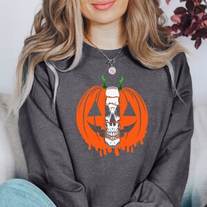 K&ouml;nnte beinhalten: Ein graues Sweatshirt mit einem Aufdruck einer K&uuml;rbis mit einem Sch&auml;del darin. Der K&uuml;rbis ist orange mit einem gr&uuml;nen Stiel und tropfendem orangefarbenem Lack.