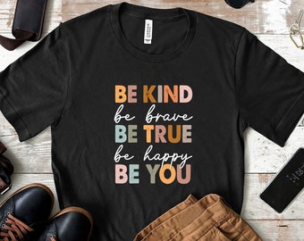 May include: Black t-shirt with a colorful graphic that reads "BE KIND be brave BE TRUE be happy BE YOU". The shirt is laid out on a white wooden surface with brown leather accessories, sunglasses, and a cell phone.