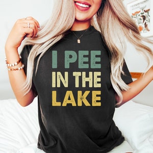 Puede incluir: Una camiseta negra con un gráfico de estilo vintage que dice "I PEE IN THE LAKE" en verde, amarillo y naranja.