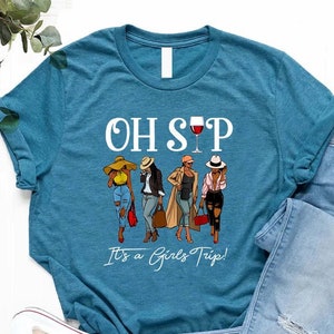 Puede incluir: Camiseta azul turquesa con un gráfico blanco de cuatro mujeres con atuendos elegantes y el texto "OH SIP It's a Girls Trip!" con una copa de vino tinto sobre el texto.