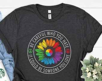 Könnte beinhalten: Ein dunkelgraues T-Shirt mit einem weißen Kreis-Motiv. Das Motiv zeigt eine Regenbogen-Sonnenblume mit Friedenszeichen und dem Text "Sei vorsichtig, wen du hasst, es könnte jemand sein, den du liebst."
