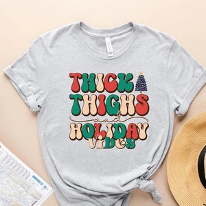 Puede incluir: Una camiseta gris con el texto "Thick Thighs and Holiday Vibes" en una fuente retro con un gr&aacute;fico de &aacute;rbol de Navidad.