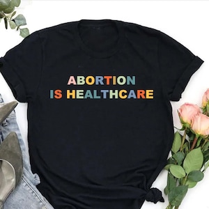 K&ouml;nnte beinhalten: Schwarzes T-Shirt mit dem Text "ABORTION IS HEALTHCARE" in Regenbogenfarben.