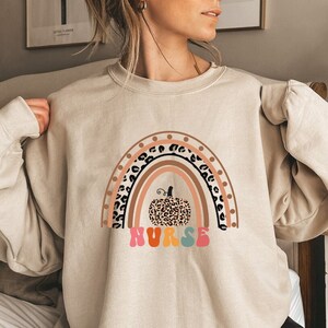 May include: A beige sweatshirt with a rainbow and a leopard print pumpkin. The rainbow has brown, pink, orange, yellow, and blue stripes. The text "NURSE" is written in rainbow colors below the pumpkin.