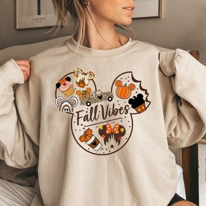 May include: Beige sweatshirt with a Mickey Mouse ear design incorporating autumn-themed illustrations. The design includes pumpkins, cupcakes, and the text "Fall Vibes". The sweatshirt is a neutral colour.