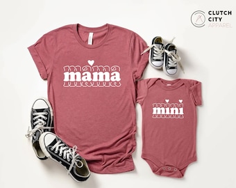 May include: A set of two shirts, one for an adult and one for a baby. The adult shirt is a maroon t-shirt with the text "mama" in white with a heart above the word. The baby shirt is a maroon bodysuit with the text "mini" in white with two hearts above the word.