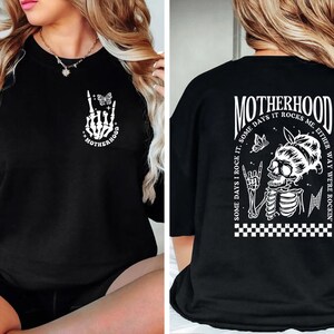May include: Black t-shirt with a white graphic print of a skeleton hand with a butterfly and the text "Motherhood" on the front. The back of the shirt has a white graphic print of a skeleton with a butterfly and the text "Motherhood Some days I rock it, some days it rocks me either way we're rockin'"