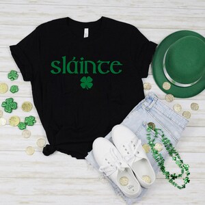 May include: Black t-shirt with the Irish phrase "Sl&aacute;inte" in green lettering and a green four-leaf clover. The shirt is laid out on a white wooden surface with a green hat, blue jeans, white trainers, and a green shamrock necklace.