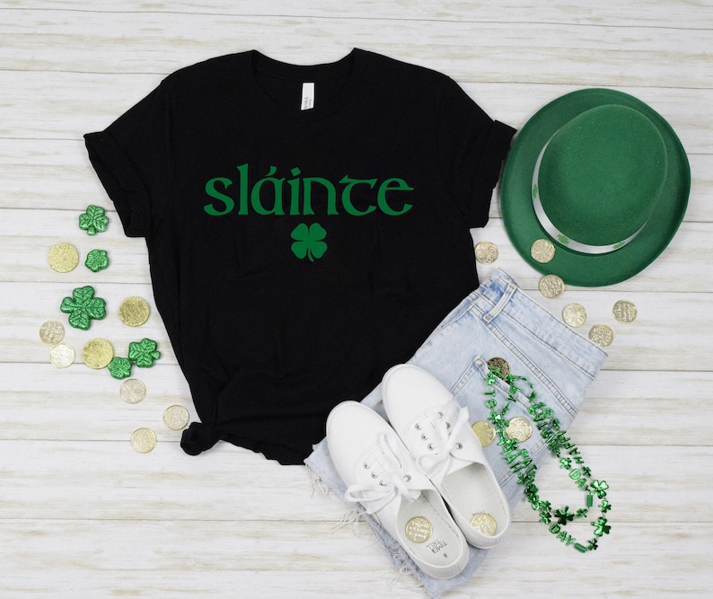 May include: Black t-shirt with the Irish phrase "Sl&aacute;inte" in green lettering and a green four-leaf clover. The shirt is laid out on a white wooden surface with a green hat, blue jeans, white trainers, and a green shamrock necklace.