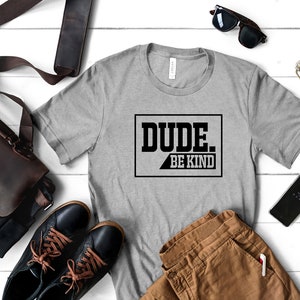 May include: A gray t-shirt with the text "DUDE. BE KIND" printed in black on the front. The shirt is laid out on a white wooden surface with a brown leather bag, a pair of brown pants, a pair of black shoes, a pair of sunglasses, and a set of keys.