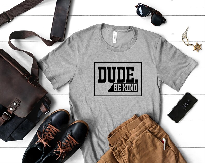 May include: A gray t-shirt with the text "DUDE. BE KIND" printed in black on the front. The shirt is laid out on a white wooden surface with a brown leather bag, a pair of brown pants, a pair of black shoes, a pair of sunglasses, and a set of keys.