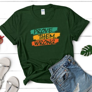 May include: A dark green t-shirt with the text "PROVE THEM WRONG" in colorful block letters. The letters are outlined in white and the background is a gradient of green, orange, and yellow.
