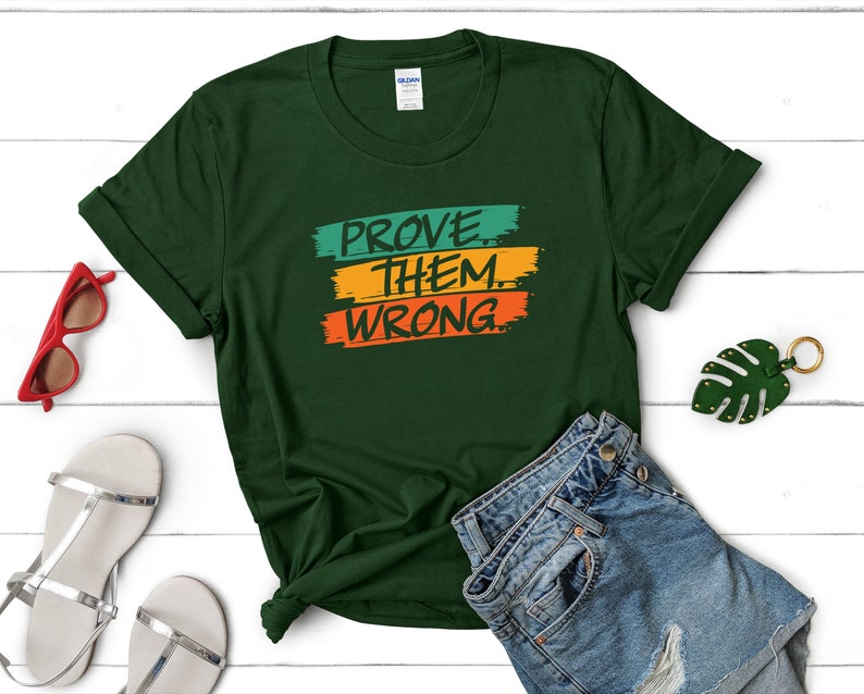 May include: A dark green t-shirt with the text "PROVE THEM WRONG" in colorful block letters. The letters are outlined in white and the background is a gradient of green, orange, and yellow.