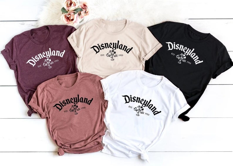 Puede incluir: Cinco camisetas de diferentes colores con el texto "Disneyland Est. 1955" y un gr&aacute;fico de cabeza de Mickey Mouse.