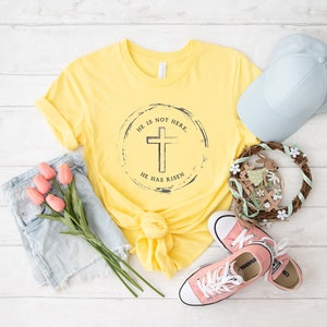 Op de afbeelding: Een geel t-shirt met de tekst "HE IS NOT HERE. HE HAS RISEN" rond een kruisafbeelding. De afbeelding bevat ook een lichtblauwe baseballpet, roze sneakers, een spijkerbroekshort en een decoratieve krans met een konijn.
