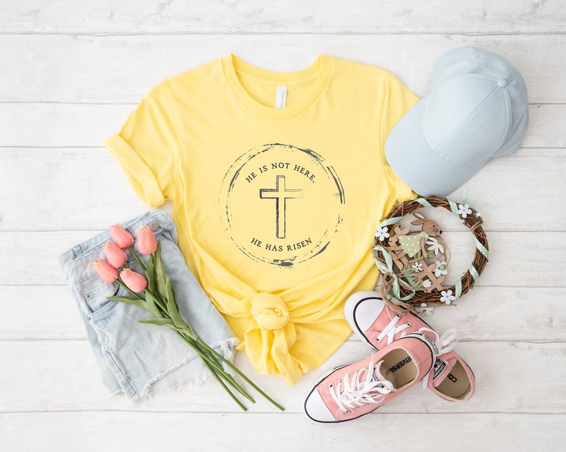 Op de afbeelding: Een geel t-shirt met de tekst "HE IS NOT HERE. HE HAS RISEN" rond een kruisafbeelding. De afbeelding bevat ook een lichtblauwe baseballpet, roze sneakers, een spijkerbroekshort en een decoratieve krans met een konijn.