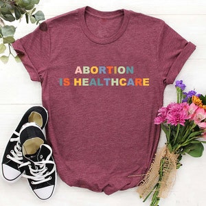 K&ouml;nnte beinhalten: Ein maroonfarbenes T-Shirt mit dem Text "ABORTION IS HEALTHCARE" in Regenbogenfarben.