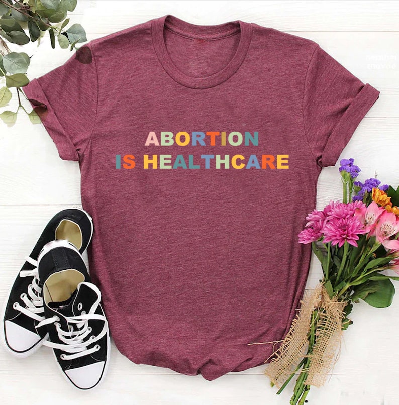 K&ouml;nnte beinhalten: Ein maroonfarbenes T-Shirt mit dem Text "ABORTION IS HEALTHCARE" in Regenbogenfarben.