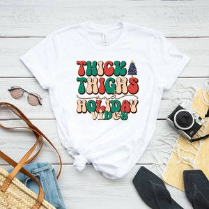 Puede incluir: Camiseta blanca con un gr&aacute;fico de estilo retro que dice "Thick Thighs and Holiday Vibes" con un gr&aacute;fico de &aacute;rbol de Navidad.
